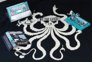 A set of turquoise cassettes in clear cases with J-cards, pictured with a band t-shirt